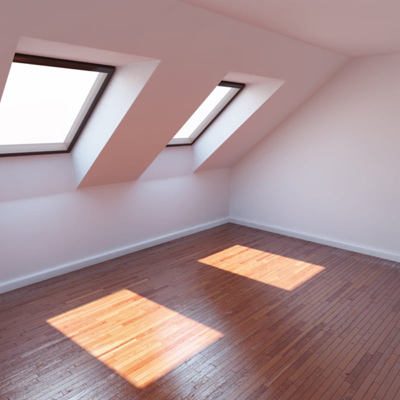 Grenier lumineux sous pente, murs blancs, plancher bois verni. Deux lucarnes projettent des carrés de lumière naturelle sur le parquet.